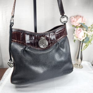 May include: Black leather shoulder bag with a brown crocodile-textured top section. The bag features a silver-toned decorative clasp and a heart-shaped charm on the strap. The bag has a single shoulder strap and a textured leather exterior.