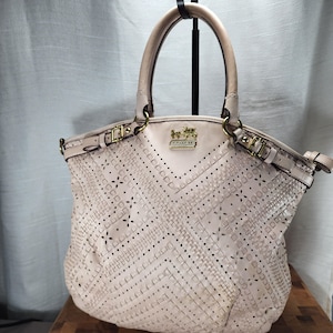 May include: A light pink leather tote bag featuring intricate laser-cut floral and geometric patterns across the front. The bag includes two top handles, gold-tone metal hardware, and a gold Coach logo plaque with a horse and carriage emblem.