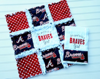May include: A white and red polka dot fabric baby quilt with a blue and white fabric square in the center. The center square has the text "It's good to be a BRAVES Girl" in red and white. The quilt is surrounded by a white ruffled border. A white washcloth with the same text is included.