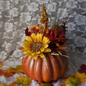 May include: A brown pumpkin with a stem and a floral arrangement inside. The arrangement includes a yellow sunflower, red flowers, and brown branches. The pumpkin is sitting on a white lace tablecloth with fall leaves scattered around it.