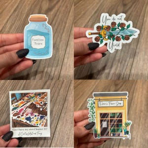 May include: Four different stickers featuring a blue glass bottle with the text "Tamlins Tears", a sword with roses and the text "You do not yield", a colorful paint splattered table with the text "Can I have my own Chapter 55? A Court of Mist and Fury", and a yellow building with the text "Elara's Flower Shop".