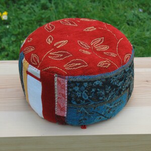 May include: A round meditation cushion with a red fabric top featuring a gold leaf pattern. The cushion is made of patchwork fabric in various colours and patterns, including blue, white, and red.