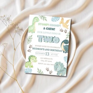 May include: A birthday invitation with a dinosaur theme. The invitation features watercolor illustrations of dinosaurs, leaves, and footprints. Text includes "STOMP! CHOMP! & CHEW! OUR LITTLE DINO IS TWO" and event details. The invitation is on a white plate.