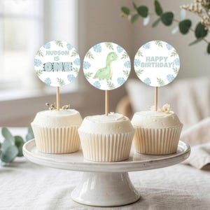 Pu&ograve; includere: Tre cupcake su un supporto bianco, ciascuno sormontato da un topper di carta circolare su uno stecchino di legno. I topper presentano un dinosauro verde chiaro, il testo "HUDSON IS ONE" e "HAPPY BIRTHDAY", decorati con motivi a foglie.