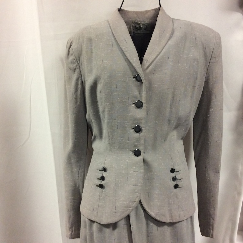 May include: A grey, textured, button-up blazer with a collar and four black buttons. The blazer has two pockets on the front with three black buttons each. The blazer is part of a matching skirt suit.