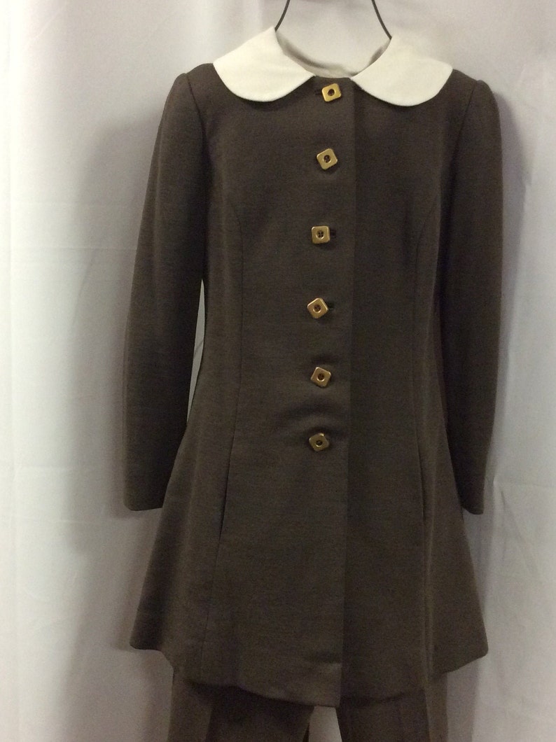 May include: A brown long-sleeved jacket with a white Peter Pan collar and gold buttons. The jacket has a slightly flared hem and is paired with matching brown pants.