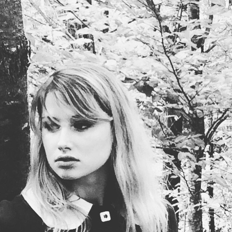 May include: A black and white portrait of a woman with long, straight hair. She is wearing a white collared shirt with a black trim. The background is a blurred image of trees.
