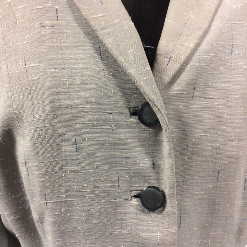 May include: A close-up of a grey blazer with a textured fabric. The blazer has two black buttons.