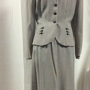 May include: A grey skirt suit with a fitted jacket and a long skirt. The jacket has a notched collar, a button fastening, and two pockets. The skirt has a straight silhouette and a waistband.
