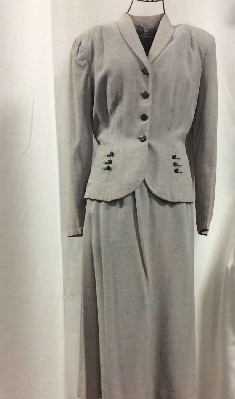 May include: A grey skirt suit with a fitted jacket and a long skirt. The jacket has a notched collar, a button fastening, and two pockets. The skirt has a straight silhouette and a waistband.