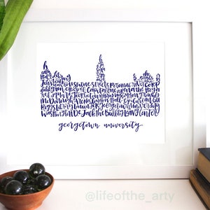 May include: A framed print featuring a navy blue silhouette of Georgetown University buildings and landmarks. The university's name is written in cursive below the skyline. The print is displayed on a white surface with decorative elements.