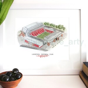 Puede incluir: Una ilustraci&oacute;n de acuarela del estadio de Anfield, hogar del Liverpool Football Club. El estadio est&aacute; representado en rojo y blanco, con c&eacute;sped verde y un marco blanco alrededor de la imagen. El texto "LIVERPOOL FOOTBALL CLUB" est&aacute; escrito en rojo debajo del estadio.