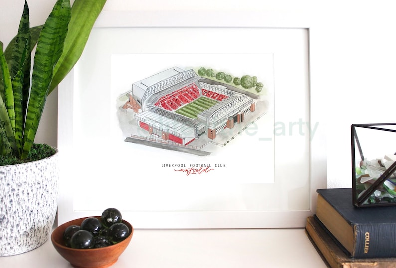 Puede incluir: Una ilustraci&oacute;n de acuarela del estadio de Anfield, hogar del Liverpool Football Club. El estadio est&aacute; representado en rojo y blanco, con c&eacute;sped verde y un marco blanco alrededor de la imagen. El texto "LIVERPOOL FOOTBALL CLUB" est&aacute; escrito en rojo debajo del estadio.