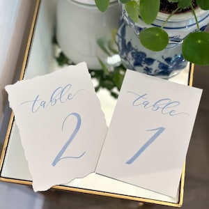 Puede incluir: Dos tarjetas de número de mesa blancas con caligrafía azul. Las tarjetas dicen "mesa 1" y "mesa 2".