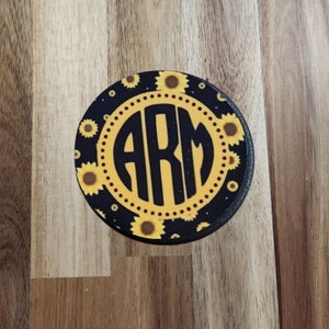 May include: A round black coaster with a white circle in the center. The circle has the letters "ARM" in black. The background of the coaster is black with a pattern of yellow sunflowers.