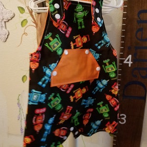May include: Black baby overalls with a colorful robot pattern. The overalls have an orange pocket and trim, and white snap closures. The garment is hanging on a white hanger.