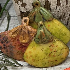 May include: A collection of handmade, colorful ceramic pendants and beads. The pendants feature floral and teardrop shapes with a mix of orange, yellow, and green hues. Some have script-like details. The beads are oval-shaped with gold-colored eyelets.