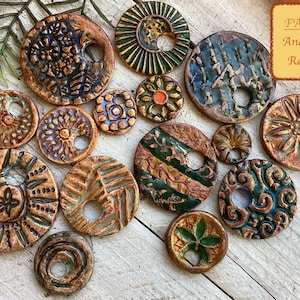 May include: A collection of faux ancient relic ceramic beads in various sizes and designs. The beads feature earthy tones of brown, green, and blue with intricate patterns and textures. Some have a central hole.