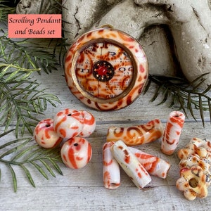 May include: A round pendant with a scrolling design and a red gemstone in the center. The pendant is surrounded by a set of white and orange beads with scrolling designs.