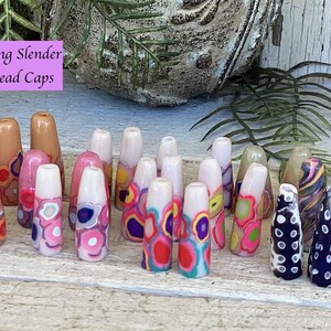 May include: A collection of 20 long, slender bead caps with colorful, abstract designs. The bead caps are made of polymer clay and have a variety of colors, including pink, white, brown, black, and blue. The bead caps are arranged in a row on a wooden surface. "Long Slender Bead Caps" is written in pink on a white background.