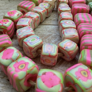 May include: A close-up of a string of cube-shaped beads in pink, green, and white. The beads feature floral and striped designs. They are strung on a natural cord, suitable for crafting jewellery or other decorative items.