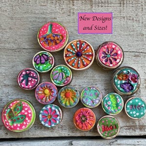 Op de afbeelding: Een verzameling kleurrijke, handgeschilderde decoratieve knopen met verschillende bloemen- en abstracte ontwerpen. De knopen zijn in tinten roze, oranje, groen en blauw. De tekst "New Designs and Sizes!" is zichtbaar.