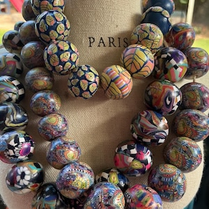 May include: A multi-strand statement necklace featuring numerous round beads in a variety of vibrant, multicolored patterns including floral, leaf, and abstract designs. The beads are arranged in clusters on a beige display form with the word PARIS.