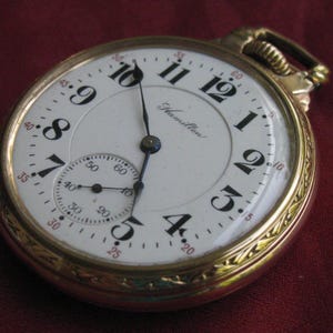 May include: A gold pocket watch with a white face, black numerals, and hands. It has a subdial and the name "Hamilton" is printed on the face. The watch is set against a burgundy background.
