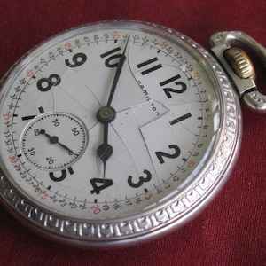 May include: A silver pocket watch with a white face and black hands. The watch has a cracked glass face and the word "Hamilton" is visible on the face.