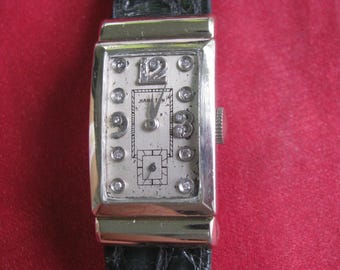 May include: Vintage rectangular wristwatch with a silver-tone case and a black leather band. The watch face is off-white with diamond accents and black numerals. The watch has a small crown and the brand name "HAMILTON" is visible.