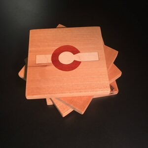 Può includere: Quattro sottobicchieri di legno impilati uno sopra l'altro. Il sottobicchiere superiore presenta un cerchio rosso e bianco con un contorno bianco, che ricorda la bandiera dello stato del Colorado.