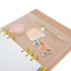 Peut inclure: Un agenda rose et or avec une pochette en plastique transparente sur le devant. La pochette contient un stylo et des autocollants avec les mots "Radiate Jesus", "Be Kind" et un arc-en-ciel.