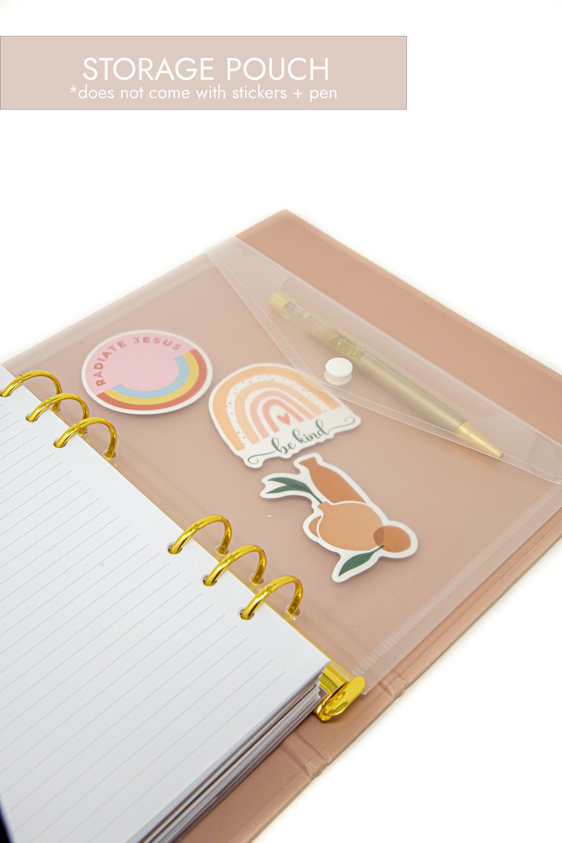 Peut inclure: Un agenda rose et or avec une pochette en plastique transparente sur le devant. La pochette contient un stylo et des autocollants avec les mots "Radiate Jesus", "Be Kind" et un arc-en-ciel.