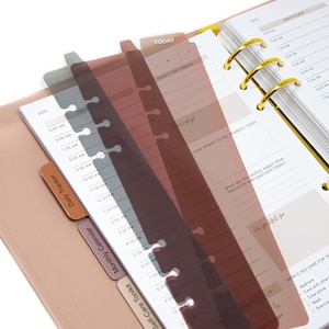 Peut inclure: Trois marque-pages translucides dans des tons de brun, gris et rose. Les marque-pages sont con&ccedil;us pour un planificateur &agrave; reliure &agrave; anneaux et comportent plusieurs trous sur le c&ocirc;t&eacute;. Les marque-pages sont &eacute;tiquet&eacute;s avec les mots "Daily Tracker", "Monthly Calendar", "Self-Care Toolkit", et "Helpful Resources".
