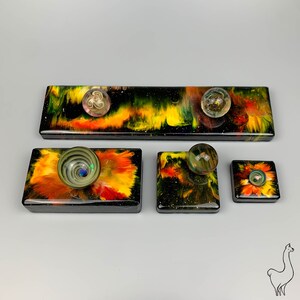 May include: Four small square resin art pieces with a black background and colorful abstract designs. Each piece features a glass marble in the center.