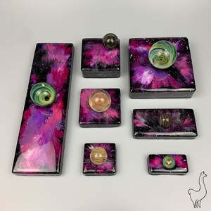 May include: A set of seven small, square, resin coasters with a galaxy-like design. Each coaster features a different colored marble in the center. The coasters are pink, purple, and black with white speckles.