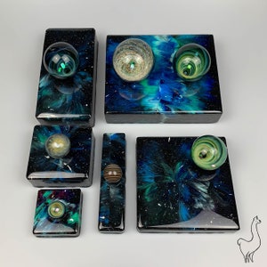 May include: A set of six small, square boxes with a galaxy-like design. Each box features a different colored glass marble with a swirling pattern. The boxes are arranged in a row, with the largest box in the center.