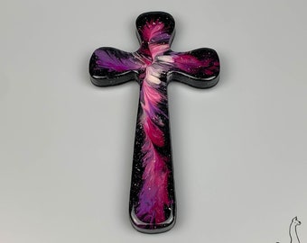 May include: A decorative cross with a black background and a vibrant, abstract design in shades of pink, purple, and white. The cross is made of a glossy material and has a smooth, rounded shape.
