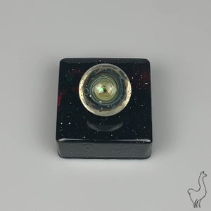 May include: A glass paperweight with a swirling, multi-colored design resembling a galaxy. The paperweight is sitting on a black square base with white speckles.