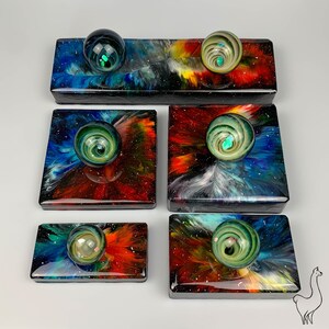May include: Five square resin coasters with a galaxy-inspired design. Each coaster features a swirling pattern of blue, red, and black with a clear glass orb in the center. The orbs have a green and blue swirl pattern.