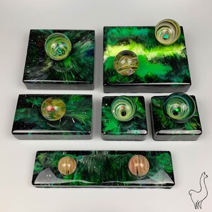 May include: Six small green and black boxes with a swirling pattern. Each box contains a marble with a different color and pattern. The boxes are arranged in two rows of three.