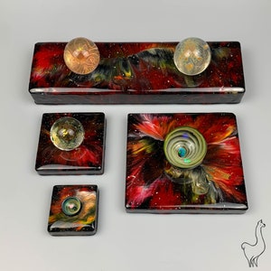 May include: Four resin art pieces with a space theme. Each piece features a swirling galaxy design with red, black, and gold accents. The pieces are arranged in a row, with the largest piece in the center. The smaller pieces are on either side of the center piece. Each piece has a clear glass orb embedded in the resin.