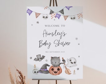 May include: A Halloween-themed baby shower sign on an easel. The sign features a banner with skulls and stars, with the text "Welcome to Ainsley's Baby Shower 3rd September 2023". The sign has illustrations of a bat, cat, pumpkin, ghost, and witch's hat.