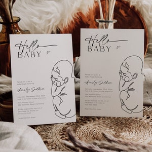 May include: Two white cards with a minimalist line drawing of a baby on each card. The text "Hello Baby" is written in a cursive font at the top of each card. The cards are on a woven brown surface.
