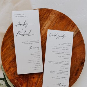 以下が含まれることがあります： 木製の表面に置かれた2つの結婚式のプログラム。プログラムは、白い背景に黒い文字で書かれています。1つは結婚式の詳細をリストし、もう1つは結婚式のパーティーをリストしています。緑の葉と白い花が前景にあります。