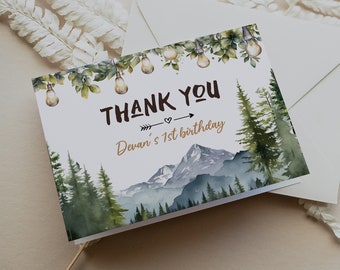 Op de afbeelding: Een witte bedankkaart met een aquarel-illustratie van een bergketen, dennenbomen en lichtslingers. De tekst "THANK YOU" is in zwart met een bruine pijl en hart. De tekst "Devan's 1st birthday" is in bruin.