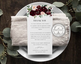 May include: A white wedding thank you card with a floral design in burgundy and blush tones, placed on a white plate with a folded linen napkin. The card reads "THANK YOU" and includes a message of gratitude.