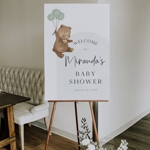 Puede incluir: Un cartel de bienvenida para un baby shower con un oso marr&oacute;n sosteniendo globos. El cartel dice "Bienvenido a Miranda's Baby Shower 12 de agosto de 2023".