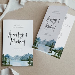 May include: Wedding invitation with a watercolor illustration of a mountain lake scene. The invitation reads "Welcome to the wedding of Ainsley & Michael" and includes the date "August 18, 2022" and location "Melbourne, Australia".