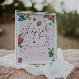 May include: A framed sign with the text "Help the busy Parents to be" in elegant script. The sign is decorated with watercolor illustrations of strawberries and blueberries on a light pink background. The frame is white.